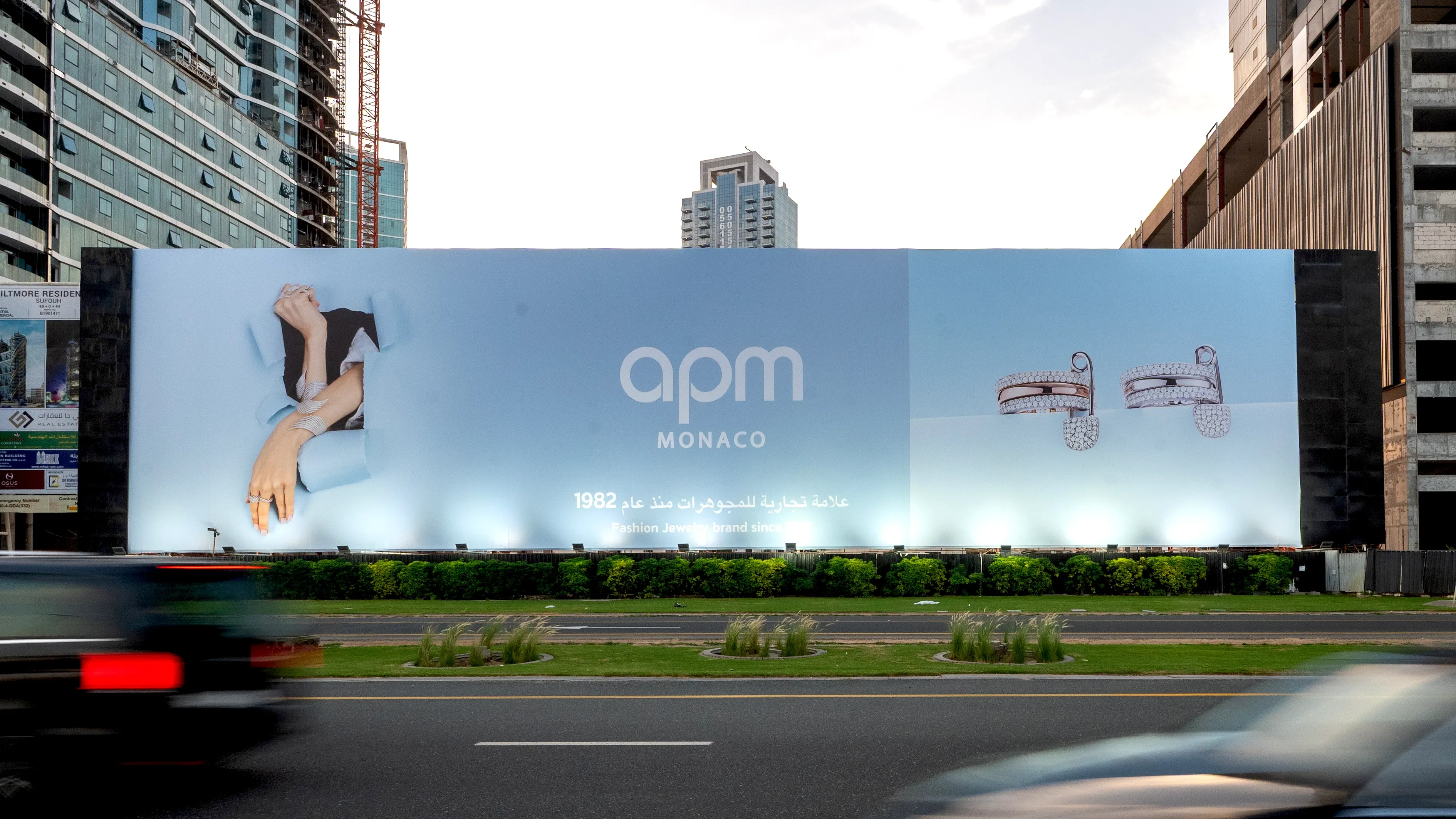 APM Monaco large-format roadside billboard advertising along a busy Dubai highway — premium static hoarding showcasing jewellery campaign with illuminated branding in an urban city setting.